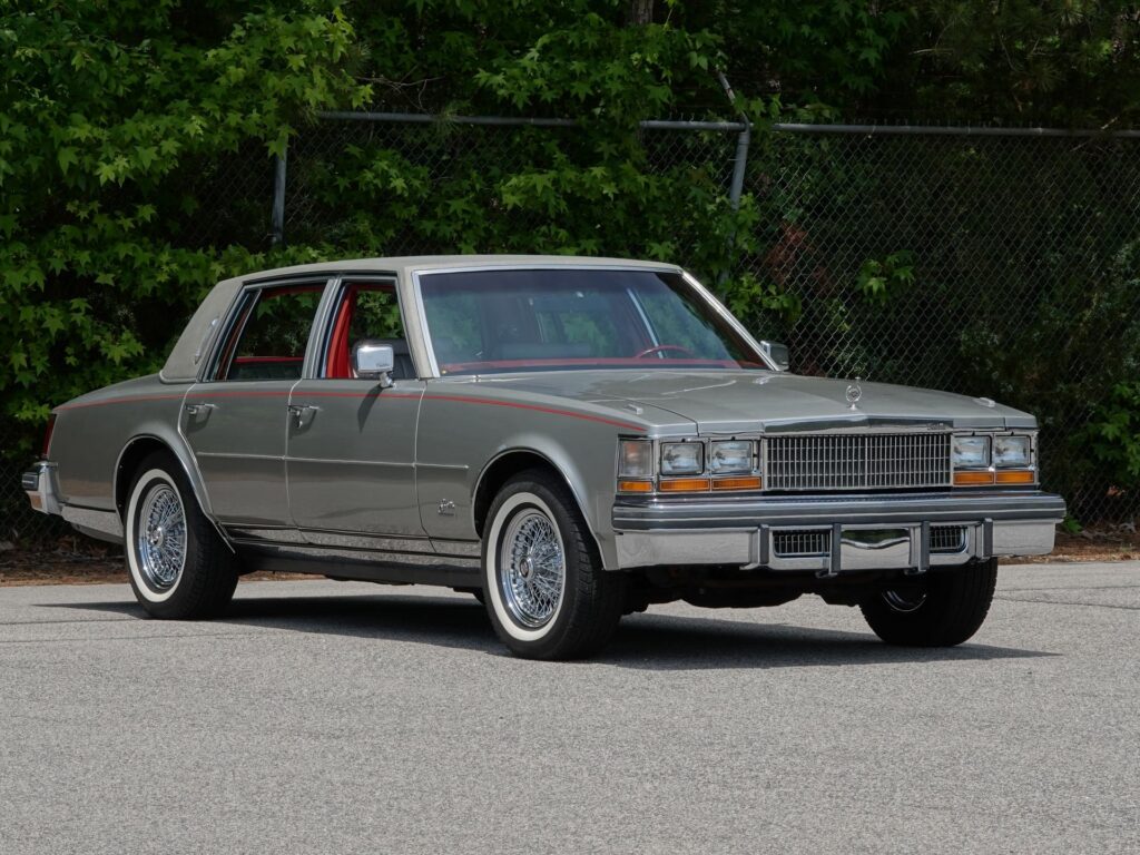 A classic 1979 Cadillac Seville parked on pavement with a chain-link fence and green foliage in the background. The car has a two-tone silver and gray paint job with a red pinstripe running along the body. It features a vinyl half-roof, wire-spoke wheels with white-wall tires, and chrome trim, grille, and bumpers. The windows are rolled down, revealing a red interior. The Cadillac hood ornament is prominently displayed on the front of the hood.